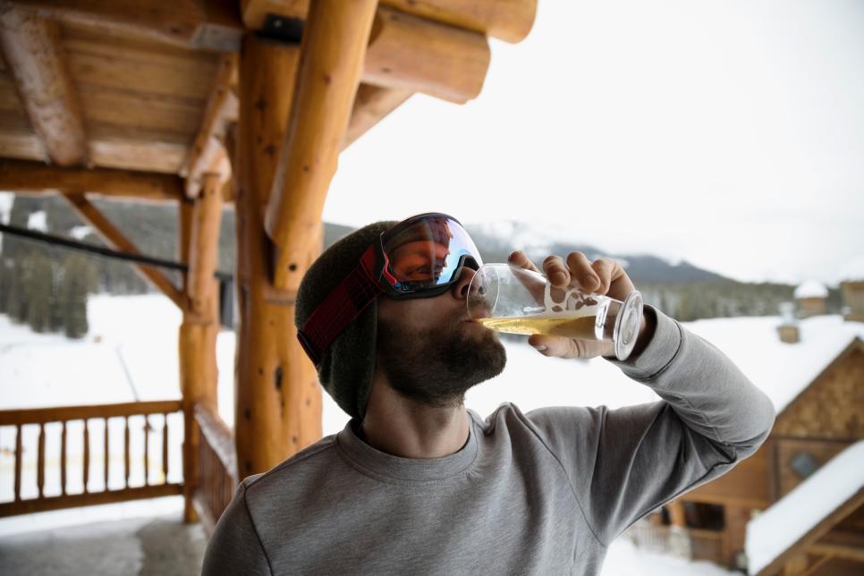Male skier in ski goggles enjoying apres-ski, drinking beer on snowy ski resort lodge balcony