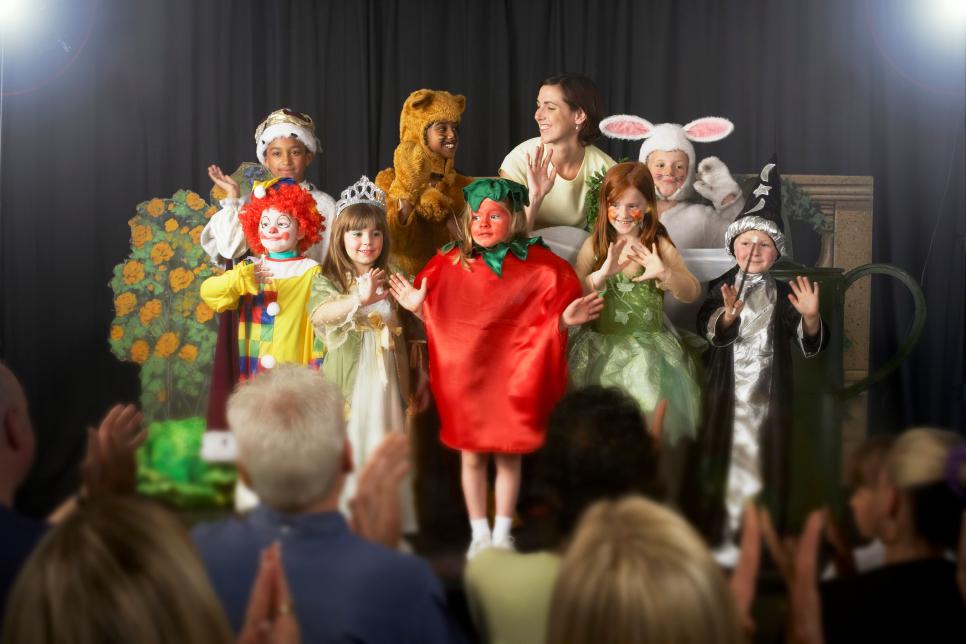 Children (4-9) wearing costumes and teacher waving on stage