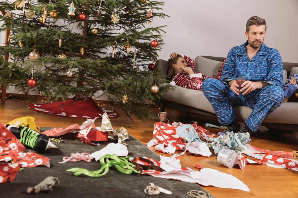 Tired couple in Christmas pyjamas looking at mess of wrapping paper