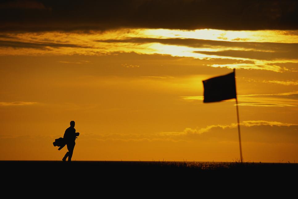 Golfer carrying clubs, walking across green at sunset, silhouette