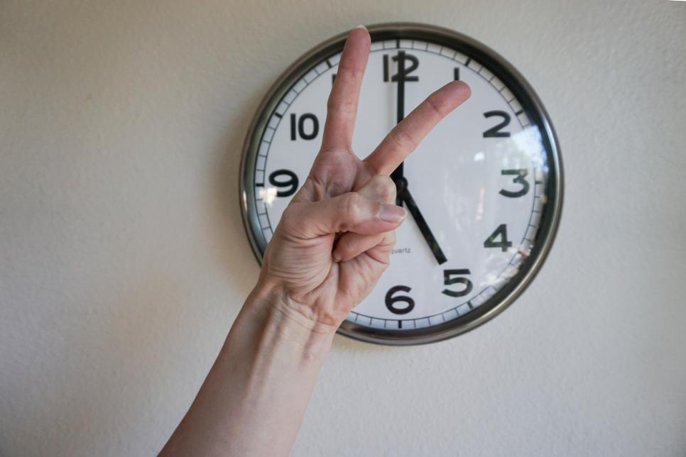 Hand showing victory sign in front of a clock, at five p.m.