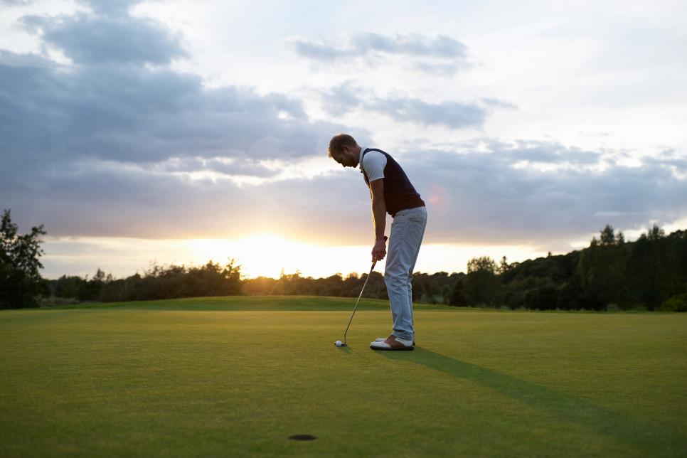 Man preparing to putt golf ball at sunset.