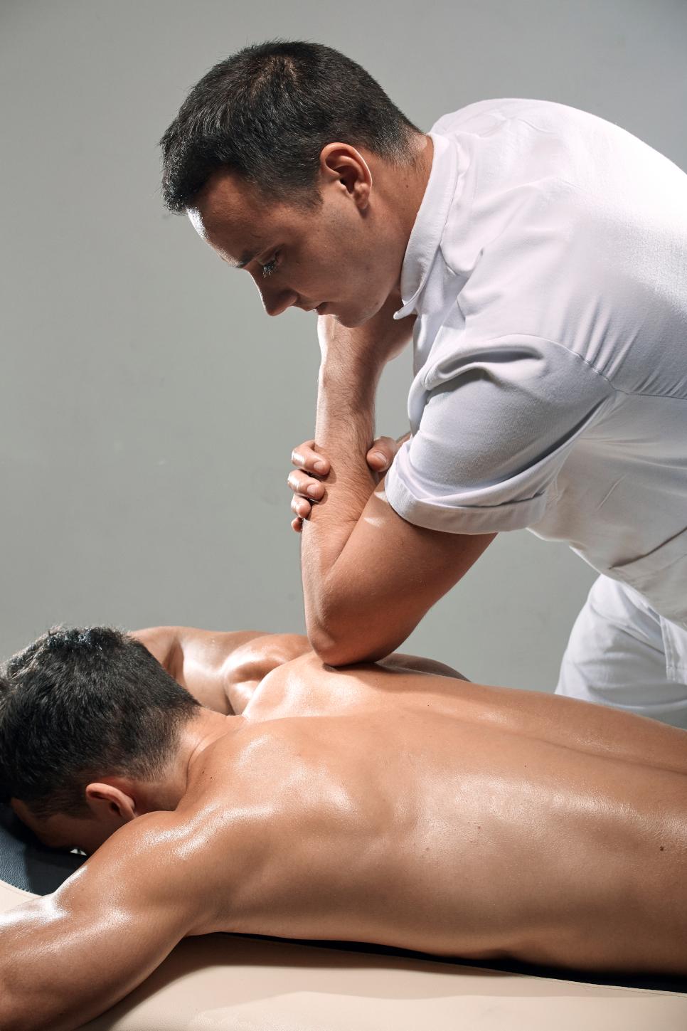 two young man, 20-29 years old, sports physiotherapy indoors in studio, photo shoot. Physiotherapist massaging muscular patient back with his elbow.