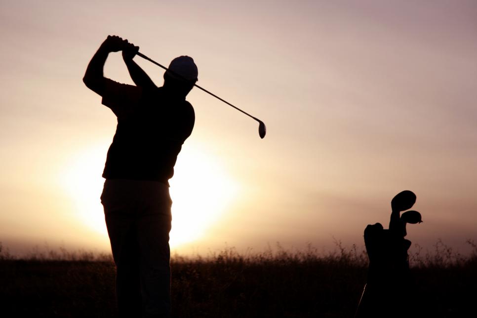 A senior golfer strikes a ball at twilight. Classic and balanced pose against the setting sun. Unrecognizable senior golfer from rear viewpoint. Golfer is healthy and fit and is Caucasian male in his 50s. One man only in image with golf equipment on beautiful links golf course in Scotland.