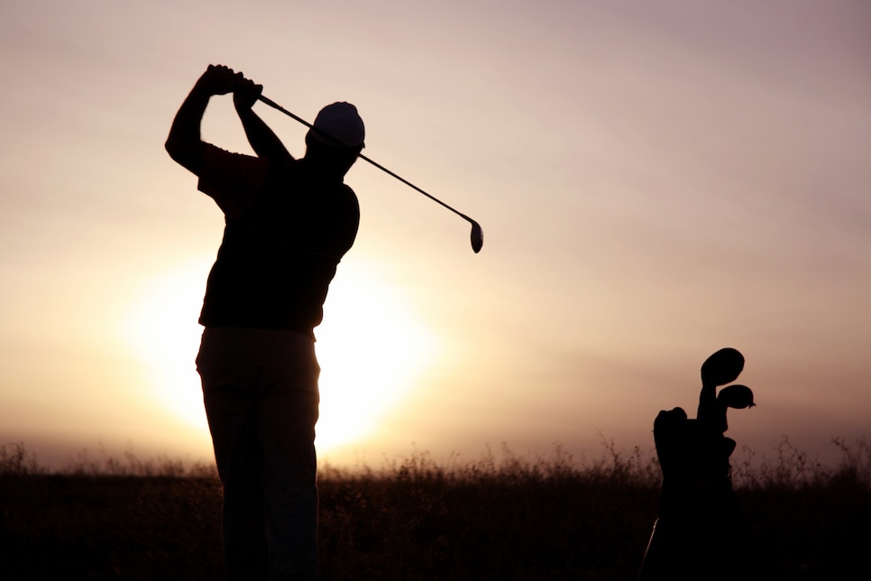 A senior golfer strikes a ball at twilight. Classic and balanced pose against the setting sun. Unrecognizable senior golfer from rear viewpoint. Golfer is healthy and fit and is Caucasian male in his 50s. One man only in image with golf equipment on beautiful links golf course in Scotland.