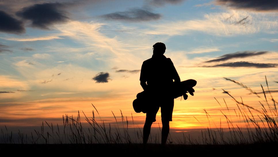 Back view of a male golfer in silhouette on a vast, windswept links course. Male golfer is a senior and is unrecognizable. Classic golf atmosphere with fescue grass, beautiful sky, and golfer walking and carrying his golf bag.