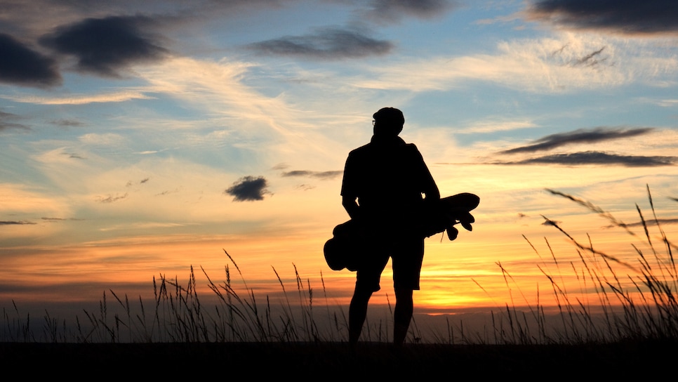 Back view of a male golfer in silhouette on a vast, windswept links course. Male golfer is a senior and is unrecognizable. Classic golf atmosphere with fescue grass, beautiful sky, and golfer walking and carrying his golf bag.