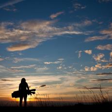 A woman in her 30s swinging a golf club against a brilliant summer sunset sky. Back view. Golf bag. Unrecognizable model. Attractive caucasian.