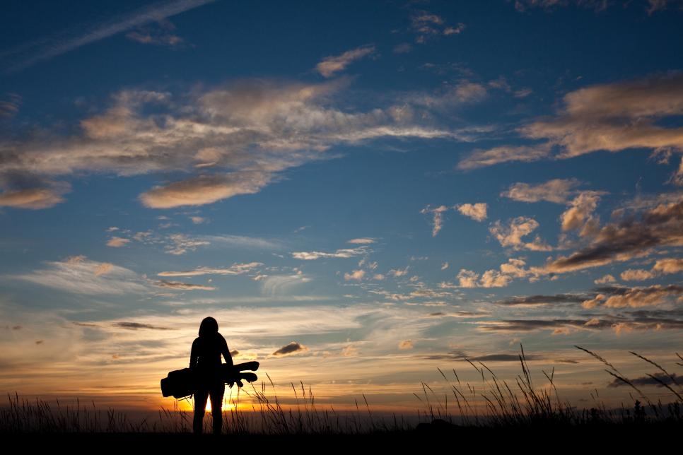 A woman in her 30s swinging a golf club against a brilliant summer sunset sky. Back view. Golf bag. Unrecognizable model. Attractive caucasian.