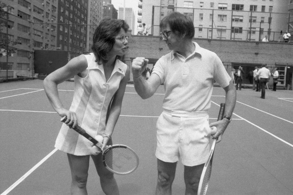 Wimbledon champion Billie Jean King tactfully holds down the net, so that 55-year-old Bobby Riggs can easily clear it during meeting at an east side tennis club here July 11th. Returning in triumph from London, Mrs. King will meet Riggs in a $100,000 winner-take-all tennis match, it was announced July 11th.